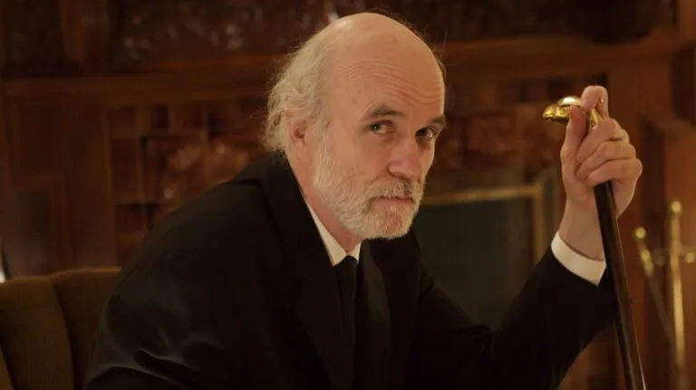 Tom Noonan jako pan Ulman v hororu The House of the Devil — herec s holí hledí přímo do kamery