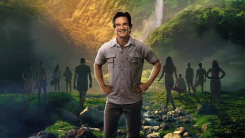 Moderátor reality show Survivor Jeff Probst na pozadí tropického prostředí s vodopádem a soutěžícími