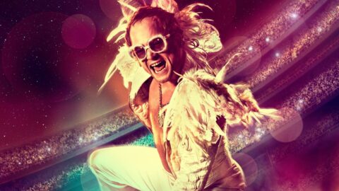 Taron Egerton jako Elton John ve filmu Rocketman, rozesmátý na koncertním pódiu v peřím kostýmu