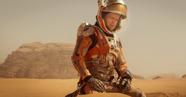Astronaut Mark Watney v oranžovo-bílém skafandru s otevřeným hledím sedí v rudé marsovské poušti ve filmu Marťan