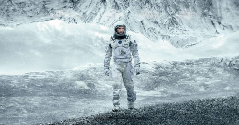 Astronaut v bílém skafandru kráčí po zamrzlé planetě se zasněženými horami v pozadí – záběr z filmu Interstellar