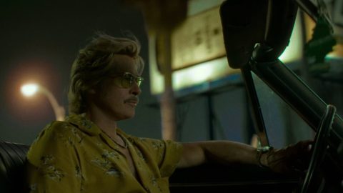 Cliff Booth ve žluté havajské košili a brýlích sedí v kabrioletu u volantu, v pozadí neonové značky