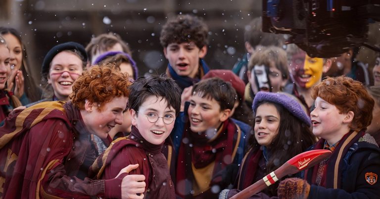 Harry s kulatými brýlemi mezi nadšenými spolužáky v nebelvírských barvách, v popředí koště Nimbus, záběr ze seriálu Harry Potter od HBO