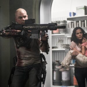 Matt Damon jako Max míří zbraní ve vesmírné stanici Elysium, v pozadí Alice Braga chrání dítě