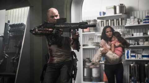Matt Damon jako Max míří zbraní ve vesmírné stanici Elysium, v pozadí Alice Braga chrání dítě